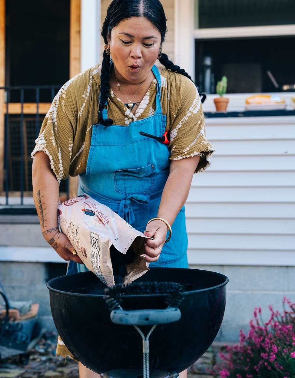 A woman outdoors prepares a grill by pouring charcoal from a bag into it, dressed in a blue apron, on a sunny day.