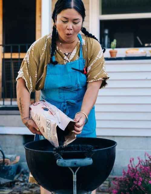 A woman outdoors prepares a grill by pouring charcoal from a bag into it, dressed in a blue apron, on a sunny day.