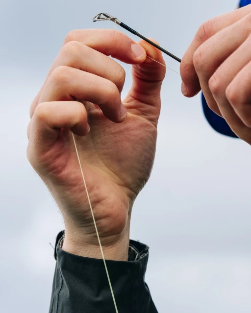 Close-up of hands tying fishing line to rod; focus on the line and rod tip against a soft, neutral sky backdrop.