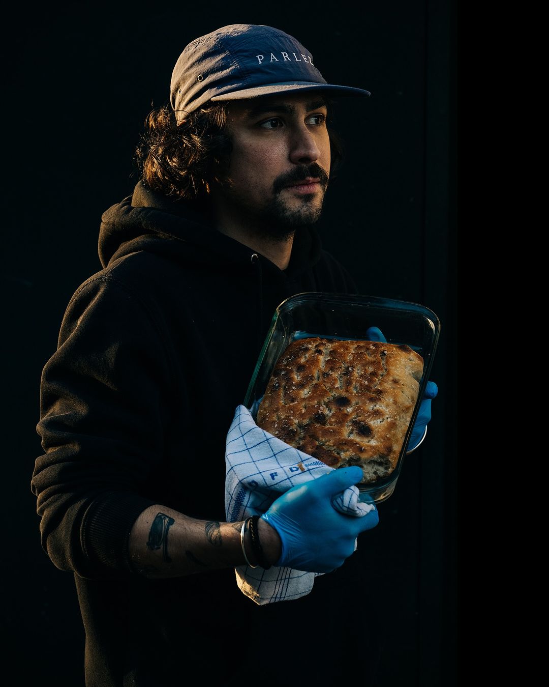 Man holding fresh baked bread in glass pan with blue gloves in dark interior. Wearing a blue cap and black hoodie.