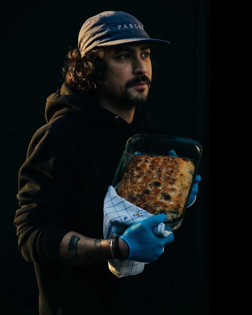 Man holding fresh baked bread in glass pan with blue gloves in dark interior. Wearing a blue cap and black hoodie.