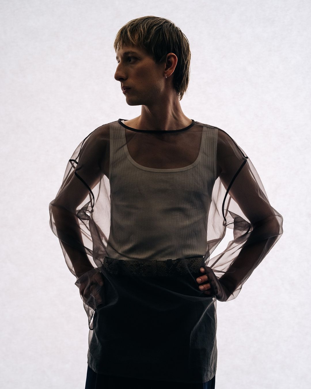 A person wearing a white tank top and a sheer, black-trimmed top stands against a bright white background in a studio setting.