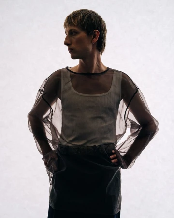 A person wearing a white tank top and a sheer, black-trimmed top stands against a bright white background in a studio setting.