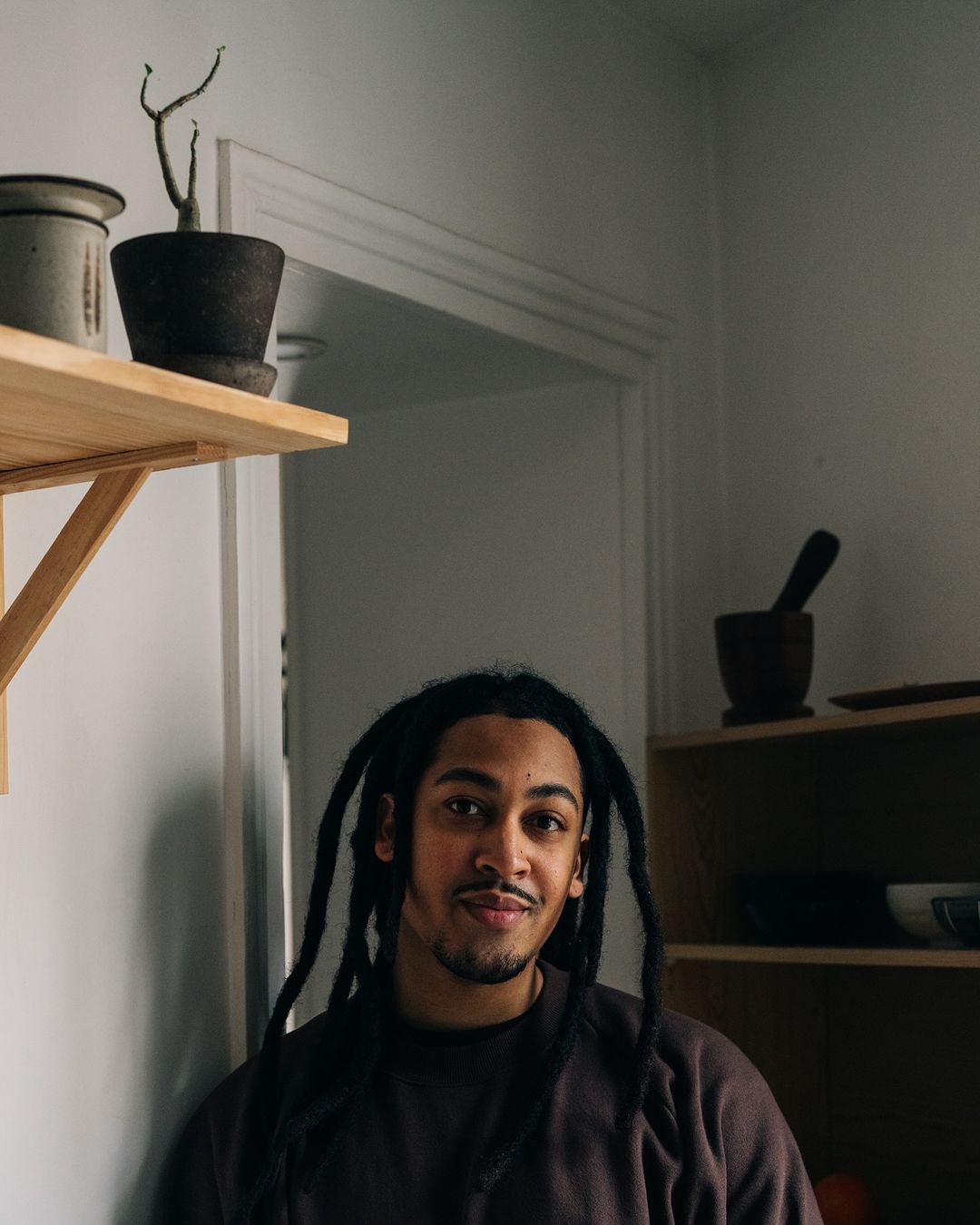 Portrait of a stylish adult man with dreadlocks, indoors near houseplants on wooden shelves against a white wall. Casual fashion.