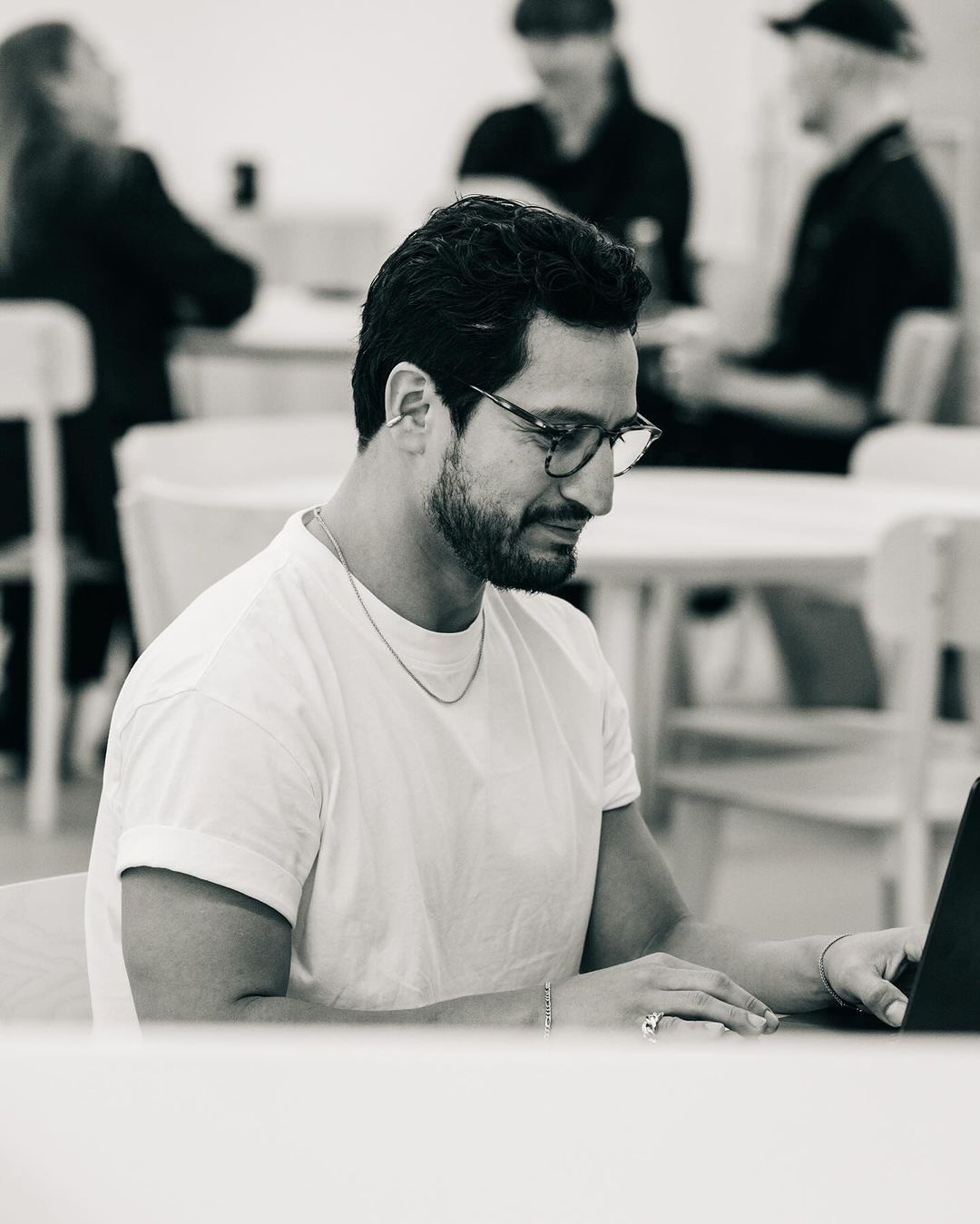 Monochrome portrait of a man with glasses working on a laptop at a table, with blurred figures in the background.