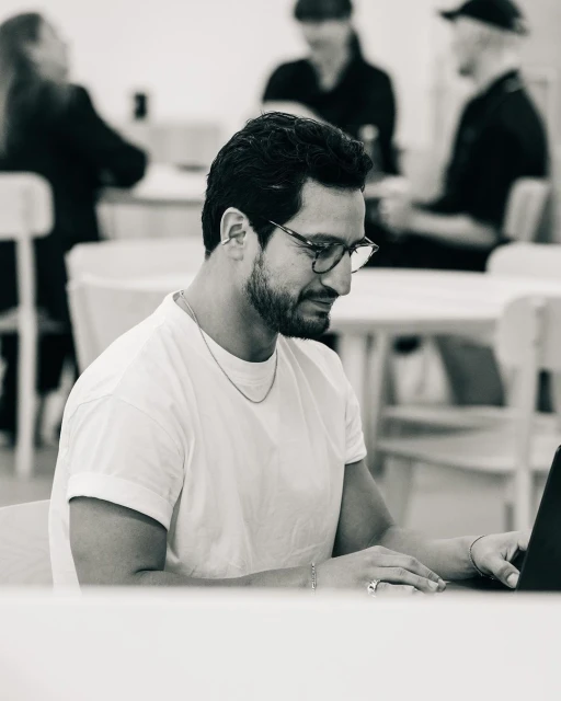 Monochrome portrait of a man with glasses working on a laptop at a table, with blurred figures in the background.
