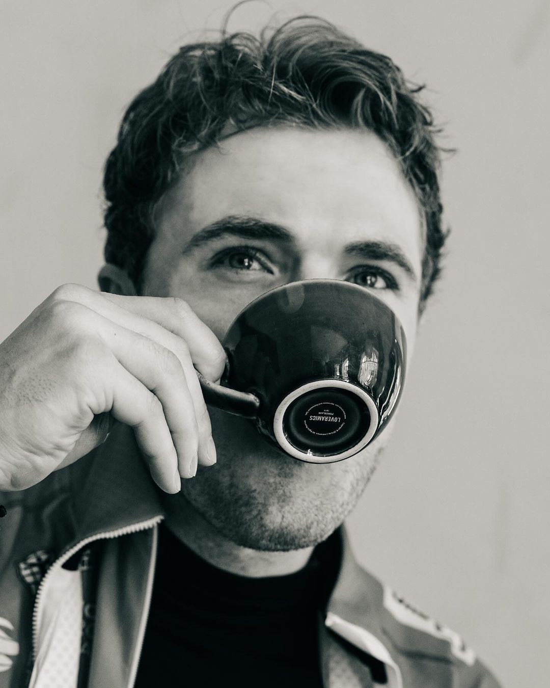 Black and white close-up of a man drinking from a dark mug, focusing on the mug's design and the man's face partially visible behind it.