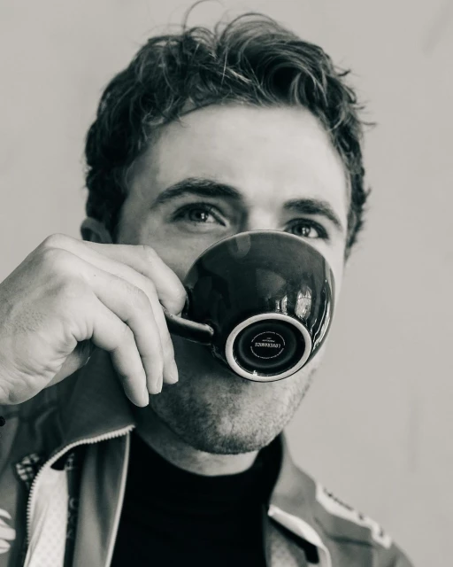 Black and white close-up of a man drinking from a dark mug, focusing on the mug's design and the man's face partially visible behind it.