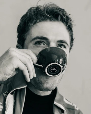 Black and white close-up of a man drinking from a dark mug, focusing on the mug's design and the man's face partially visible behind it.