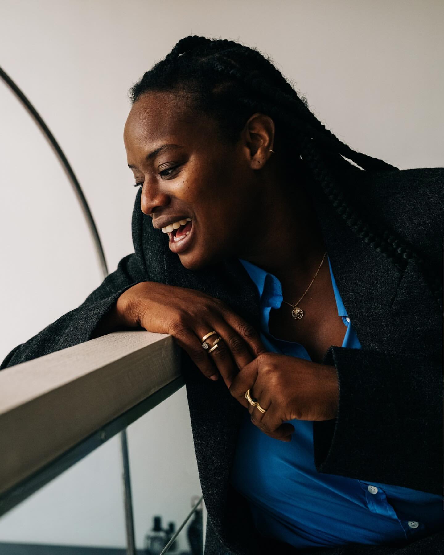 Smiling Black woman in a blue shirt and grey blazer, leaning on a white ledge. Jewelry and rings are visible. Indoor setting, soft lighting.