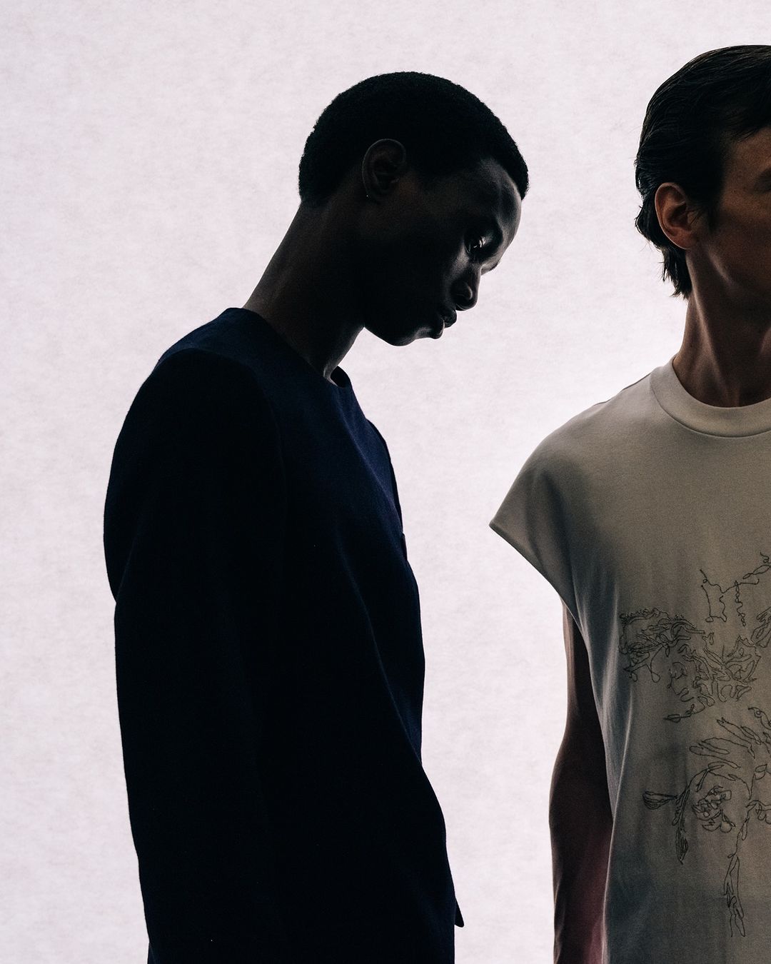 Two adult models stand against a bright white backdrop, showcasing a stark fashion photography style with minimalist aesthetic and muted tones.