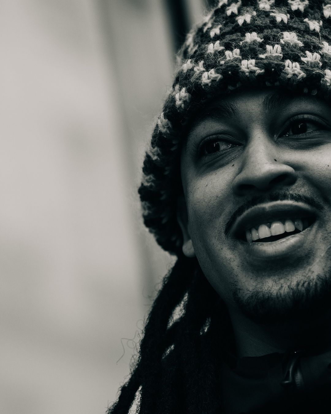 Close-up portrait in black and white of an adult man with locs and a knit hat, smiling and looking off to the side.