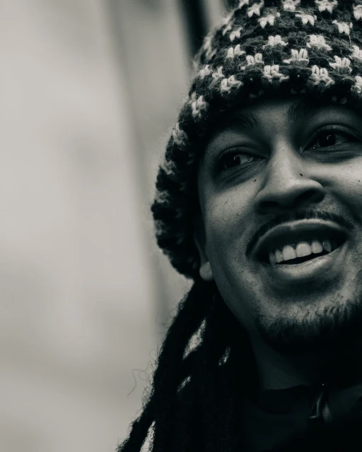 Close-up portrait in black and white of an adult man with locs and a knit hat, smiling and looking off to the side.