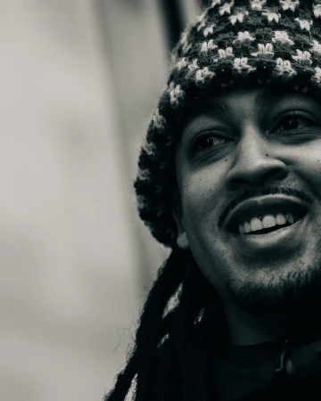Close-up portrait in black and white of an adult man with locs and a knit hat, smiling and looking off to the side.