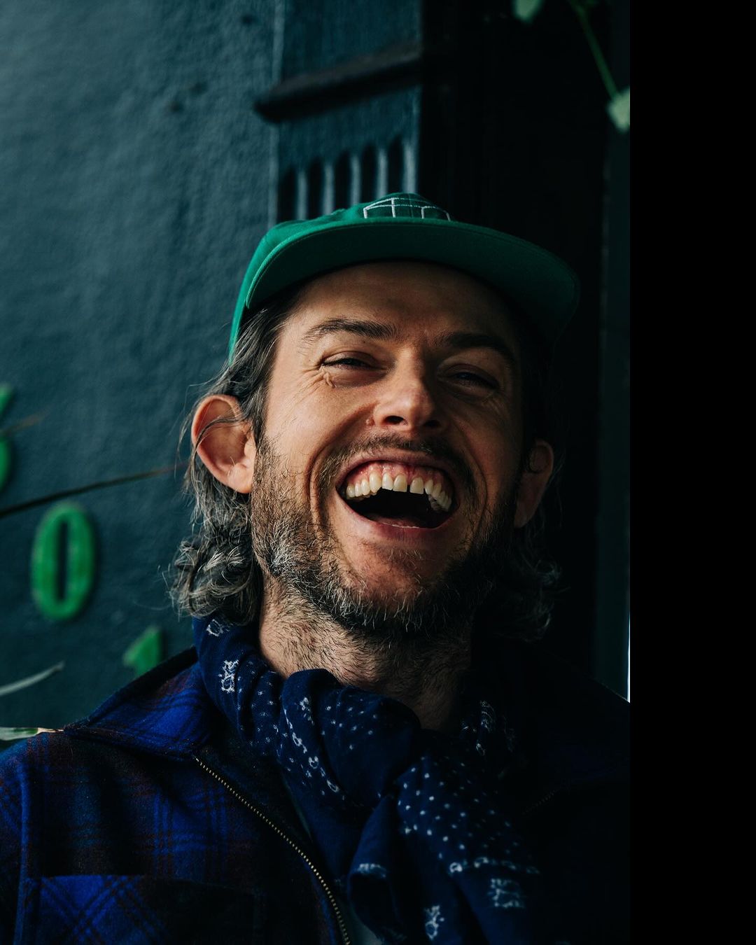 Man wearing a green cap and blue plaid jacket with a dark patterned scarf laughing outdoors in New York by Adam Katz Sinding