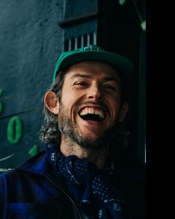 Man wearing a green cap and blue plaid jacket with a dark patterned scarf laughing outdoors in New York by Adam Katz Sinding