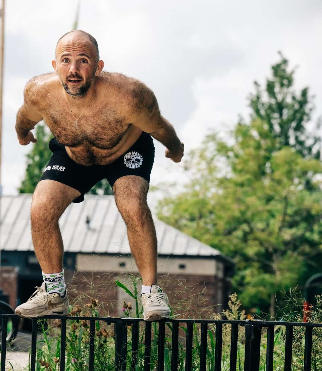 Adult male performing a fitness routine outdoors. Man is shirtless, wearing athletic shorts and sneakers, balancing on a metal rail.