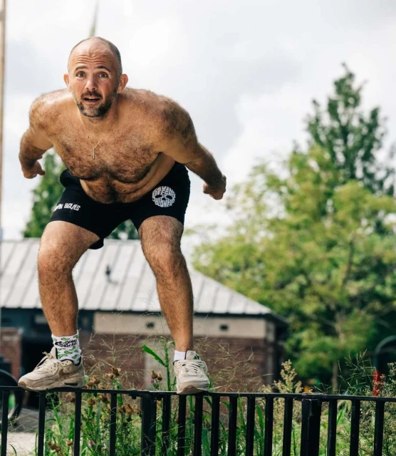 Adult male performing a fitness routine outdoors. Man is shirtless, wearing athletic shorts and sneakers, balancing on a metal rail.
