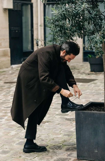 A bearded man in a long coat tying his shoelaces outside on a cobblestone street near a tree, with a building in the background.