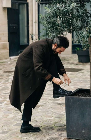 A bearded man in a long coat tying his shoelaces outside on a cobblestone street near a tree, with a building in the background.