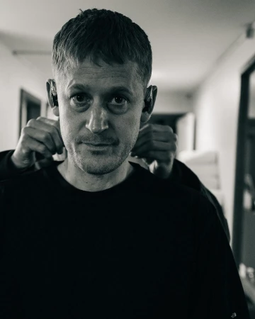 Monochrome portrait of an adult male wearing hearing aids, another person adjusting his collar in an indoor setting with muted tones.