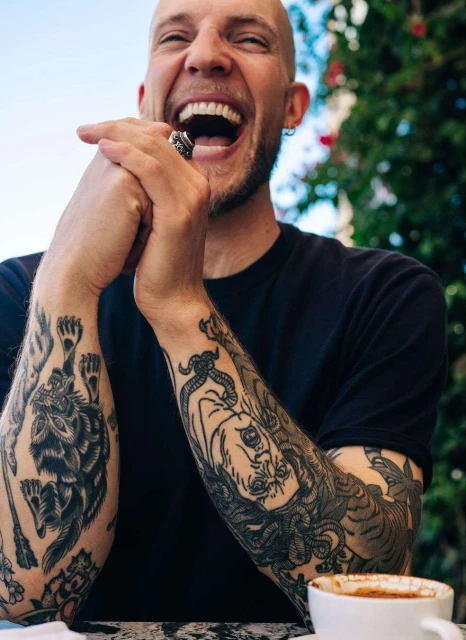 Adult man laughs in outdoor cafe. He has tattoos on his arms and is wearing a black t-shirt with plants in background.