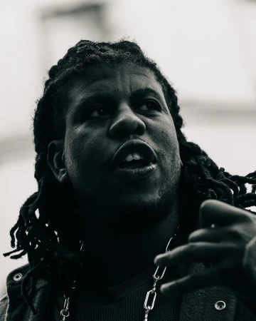 Black and white portrait of a person with locs wearing a silver chain looking up with an open mouth and gesturing with a hand.