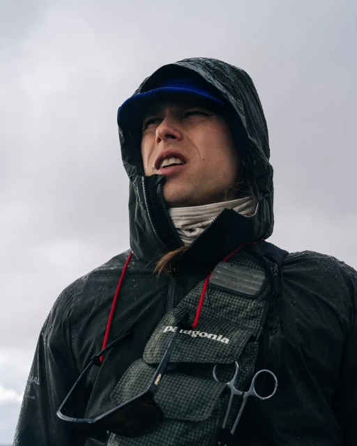 A man in a hooded black jacket and blue baseball cap looks up in an outdoor setting, wearing a fishing vest with sunglasses and scissors.