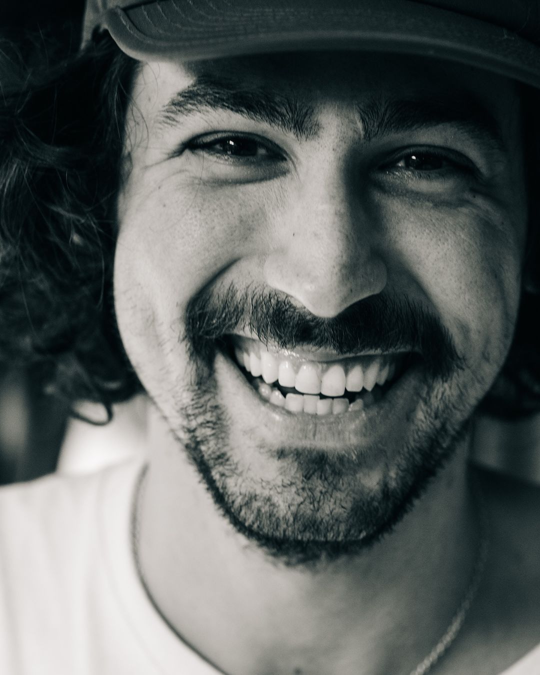 Close-up monochrome portrait of a smiling man wearing a cap and t-shirt, photographed by Adam Katz Sinding in New York.