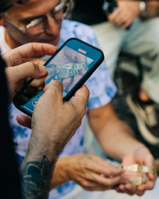 Person recording a luxury watch on a smartphone. Close-up of hands holding the phone and watch indoors, with an out-of-focus person in the background.
