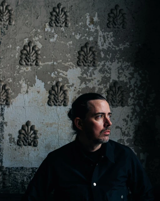 Adult male in a dark jacket poses indoors against an ornate wall with decorative plaster details and peeling paint in a moody portrait.