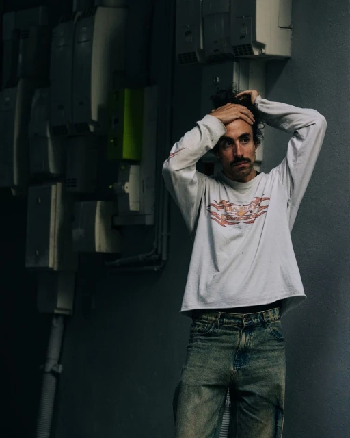 Adult male in a white long-sleeve shirt and jeans poses indoors against a background of electrical boxes and pipes. Dark and moody.