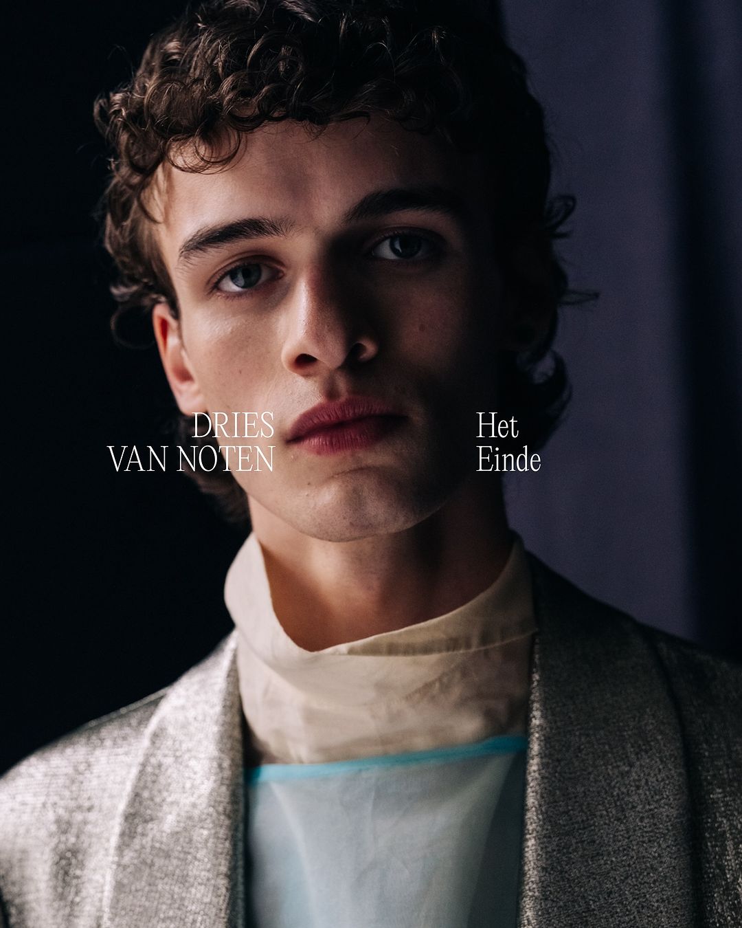 Close-up portrait of a young adult male with curly hair wearing a light-colored top and metallic silver jacket against a dark background.