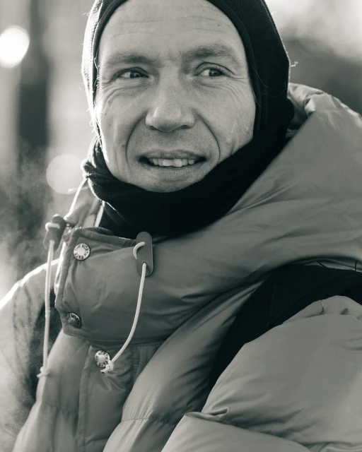 Monochrome portrait of a middle-aged man wearing a knit cap and a down jacket, vapor visible in the cold air.