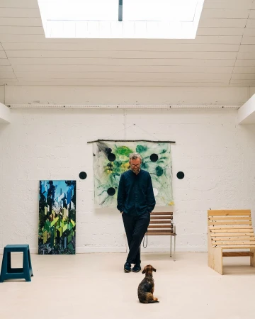 Man standing in white art studio with a small dog looking up at him, by Adam Katz Sinding