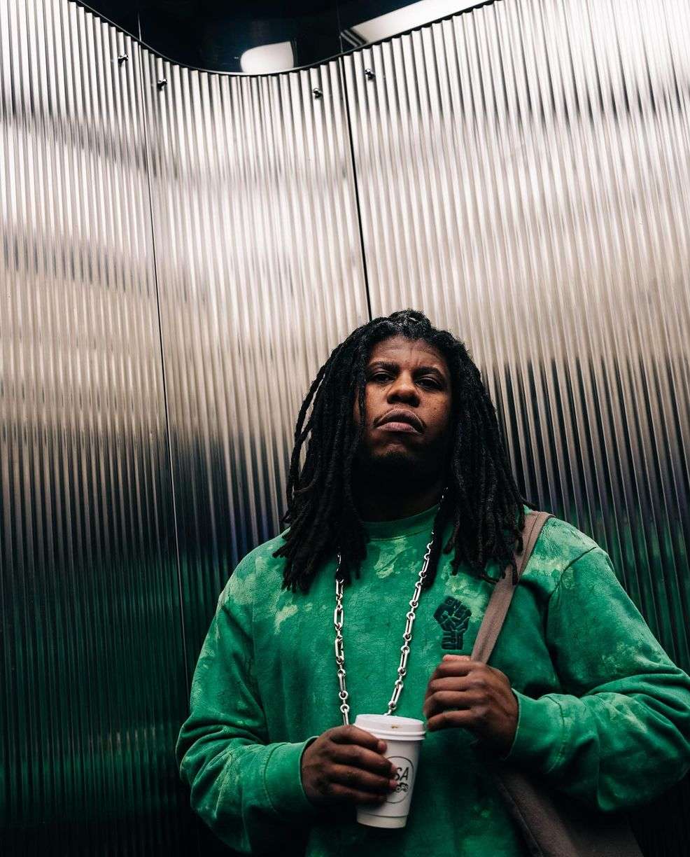 Low-angle view of an adult male with dreadlocks wearing a green tie-dye sweatshirt and holding a disposable cup in a metal elevator.