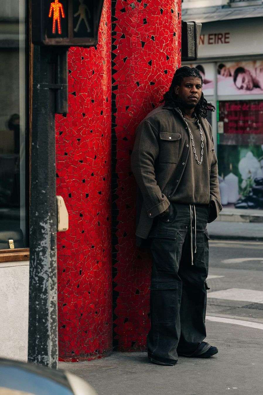 Adult male in casual attire stands next to a red mosaic-covered column on a city street, with a traffic light visible nearby.