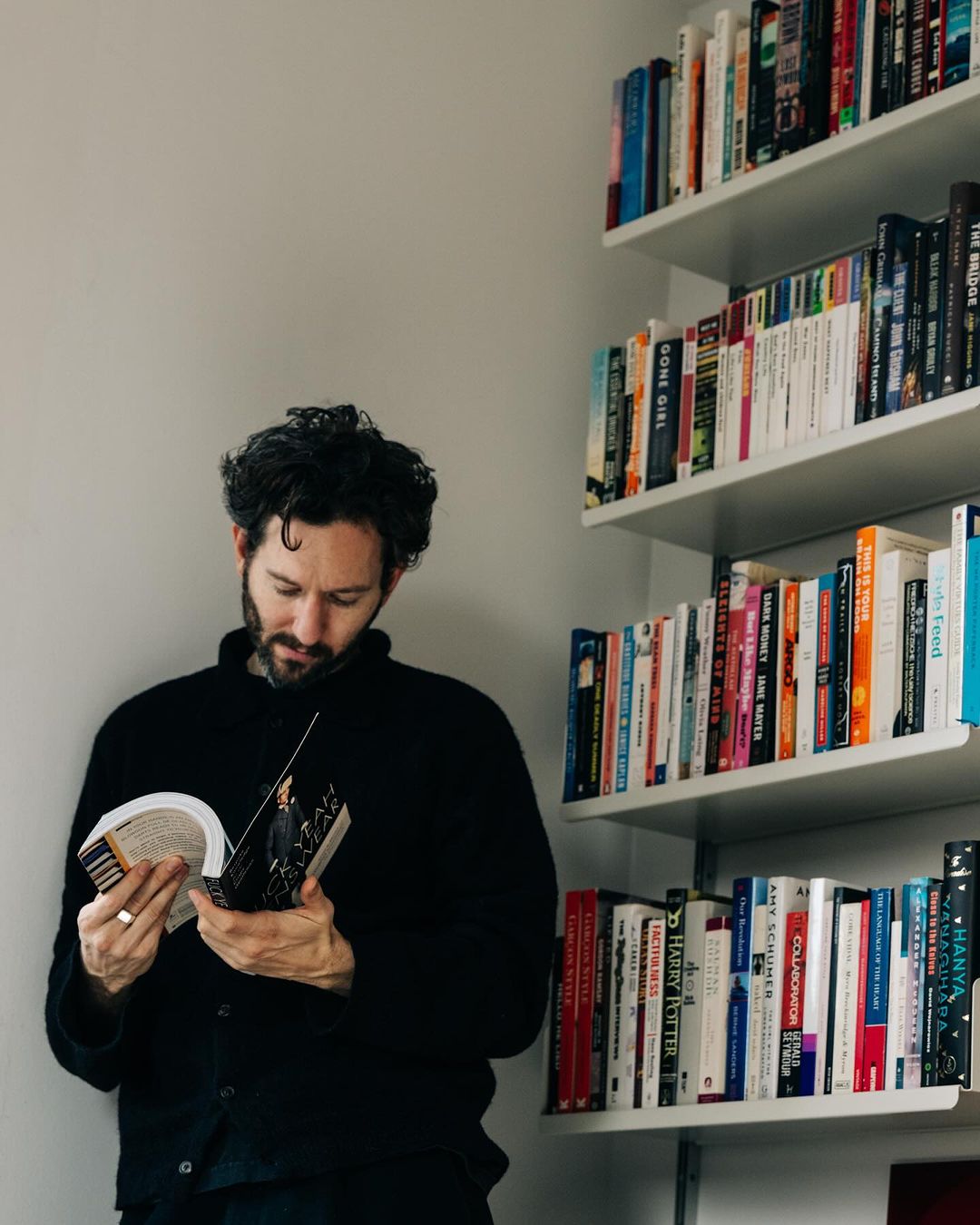 Adult man with beard reading a book indoors, next to a white bookshelf filled with books, wearing a dark cardigan.