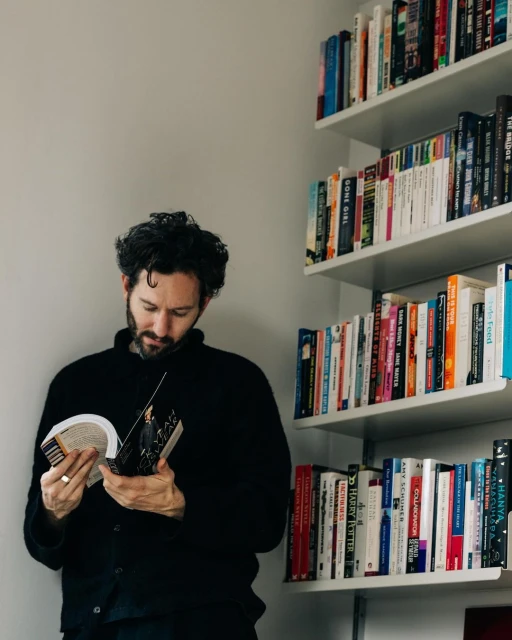 Adult man with beard reading a book indoors, next to a white bookshelf filled with books, wearing a dark cardigan.