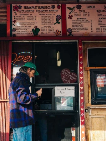 Man in a green cap stands at an outdoor food window with a colorful hand-painted menu sign, ordering food from a walk-up window.