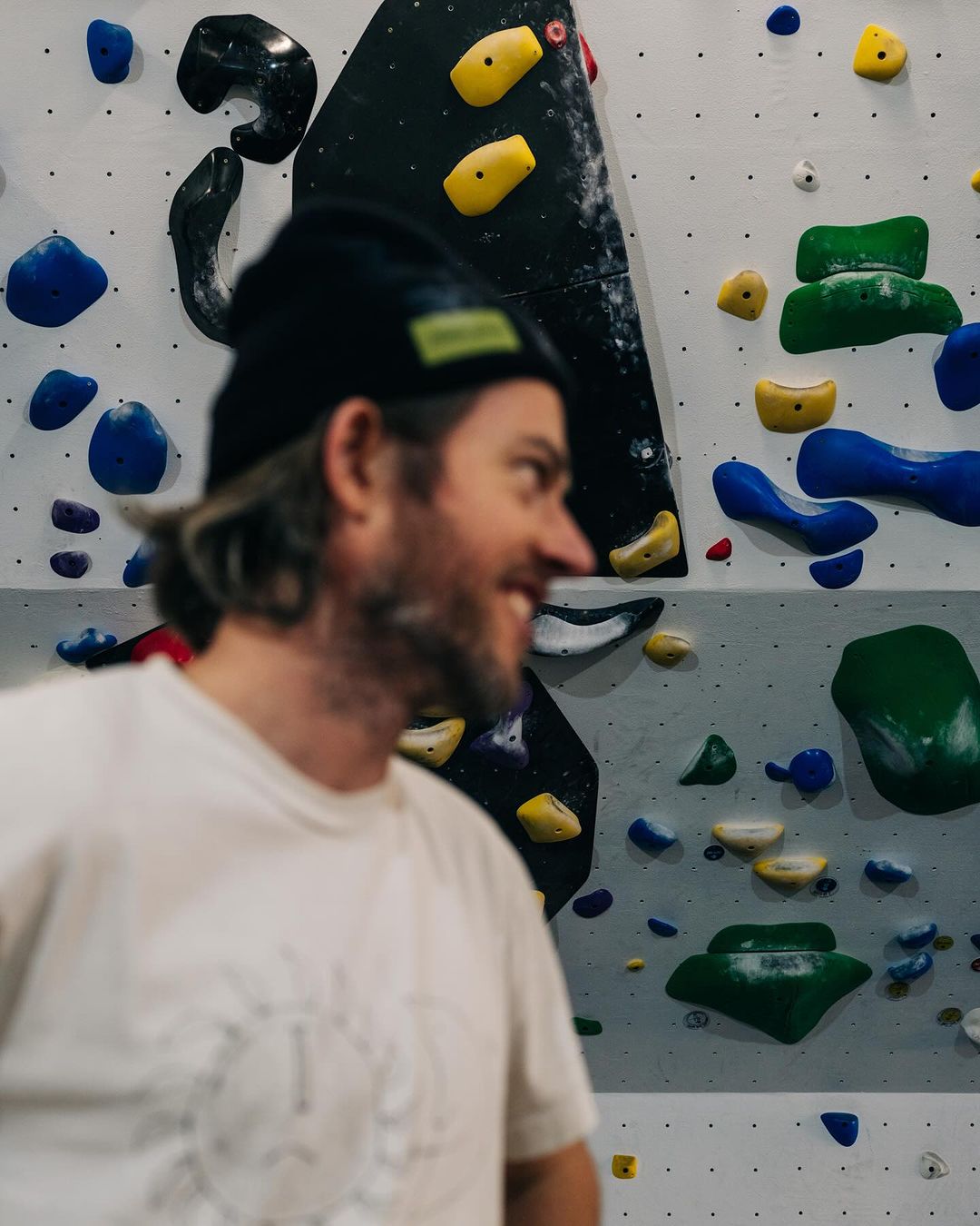Close-up of a smiling adult man with a beanie, set against a climbing wall with colorful holds in an indoor gym setting.