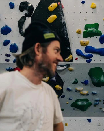 Close-up of a smiling adult man with a beanie, set against a climbing wall with colorful holds in an indoor gym setting.