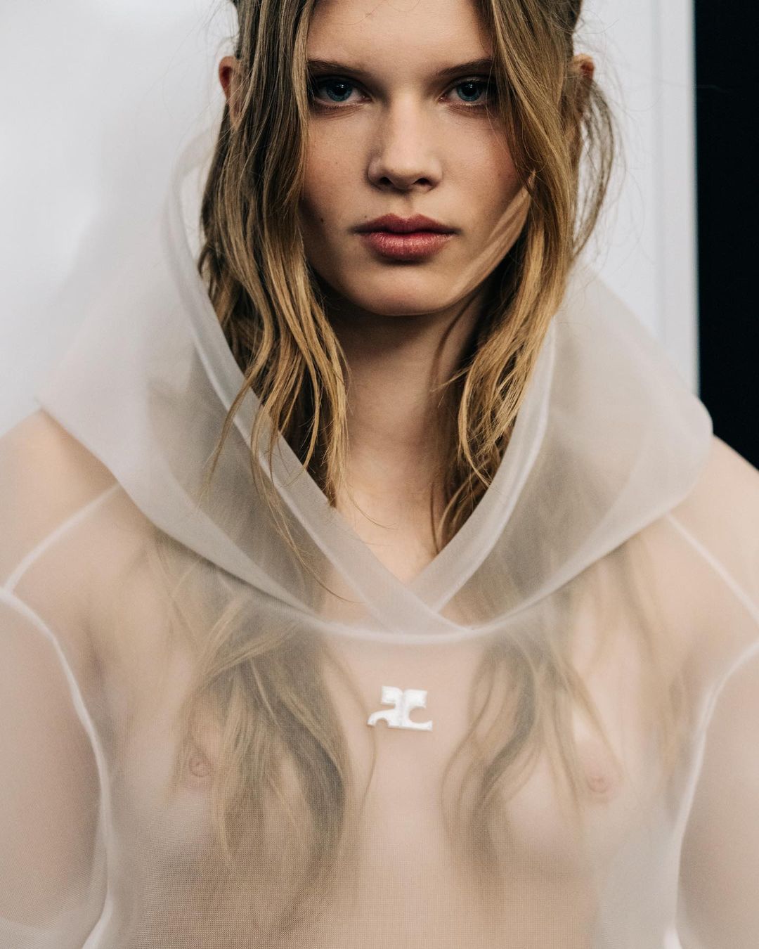 Close-up portrait of a blonde woman in a sheer hooded top with a logo, featuring a fair complexion and soft, diffused lighting.