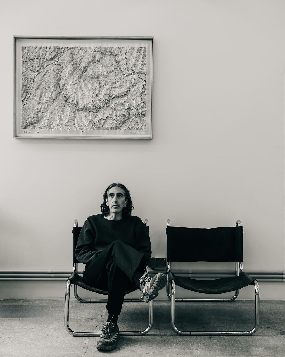 Middle-aged man with black hair sits on a black chair indoors in front of a monochrome map in a minimalist setting.