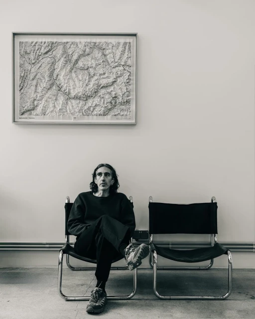Middle-aged man with black hair sits on a black chair indoors in front of a monochrome map in a minimalist setting.