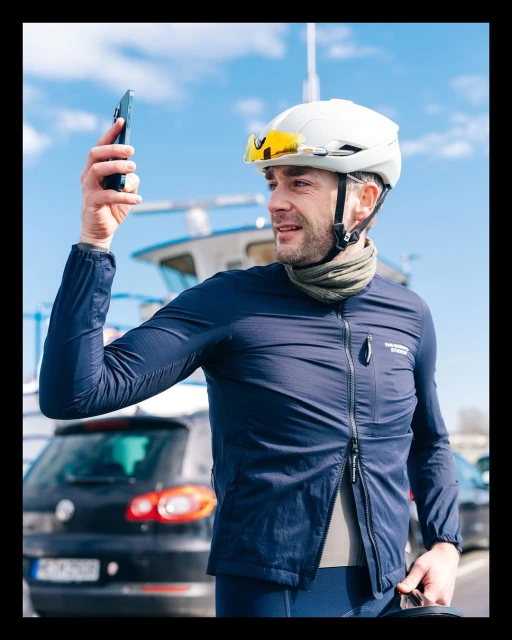 Adult man wearing a cycling helmet and jacket takes a selfie with a smartphone outdoors near a parked car and boat on the water.