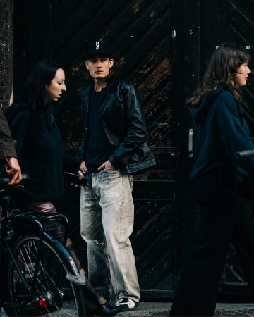 Group of people on a city street with a man in a black leather jacket and baseball cap standing in the center
