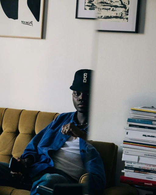 Man sits on mustard yellow sofa in blue jacket and black cap. Stack of books, framed art prints, and white walls create a cozy indoor scene.