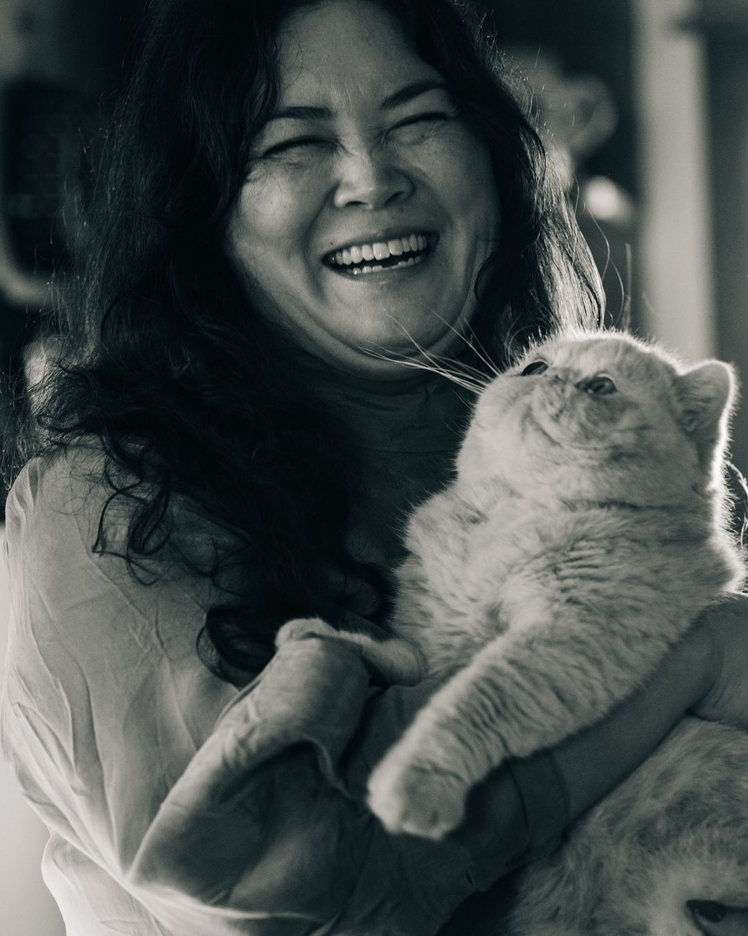 Monochrome portrait of a smiling woman holding a beige cat with closed eyes in her arms indoors, in a close-up shot.
