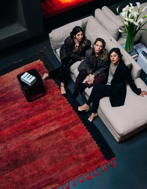 Three women sit on a beige couch in an interior space, showcasing modern clothing, a red rug, and a vase of white flowers.